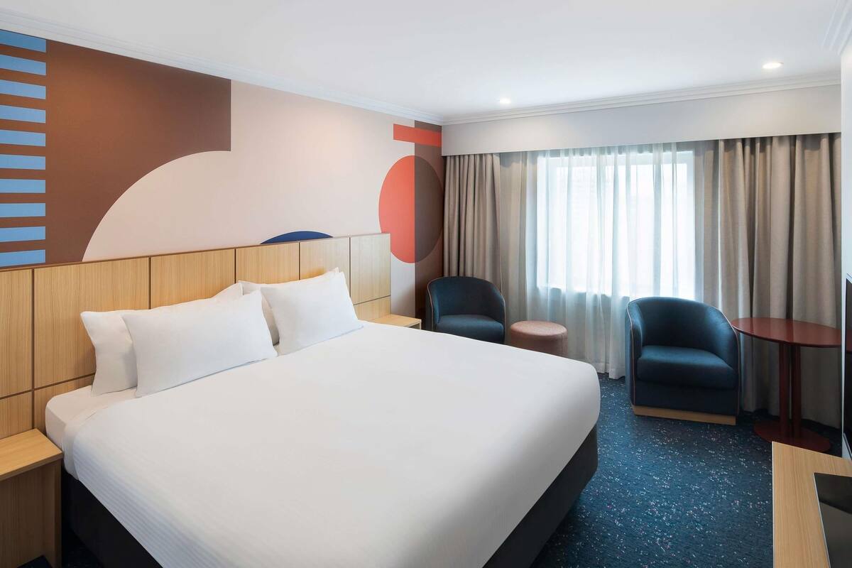 A hotel room with a large bed, two chairs, a small table, and a TV.