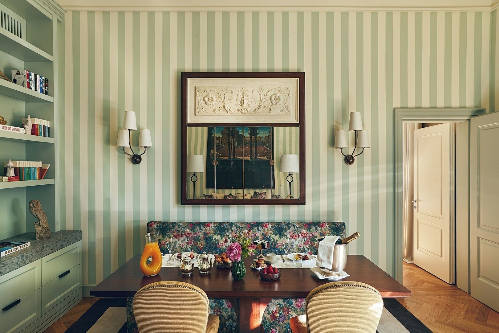 A dining area with a striped wall, a framed mirror, two wall sconces, a floral bench, and a table set for a meal.