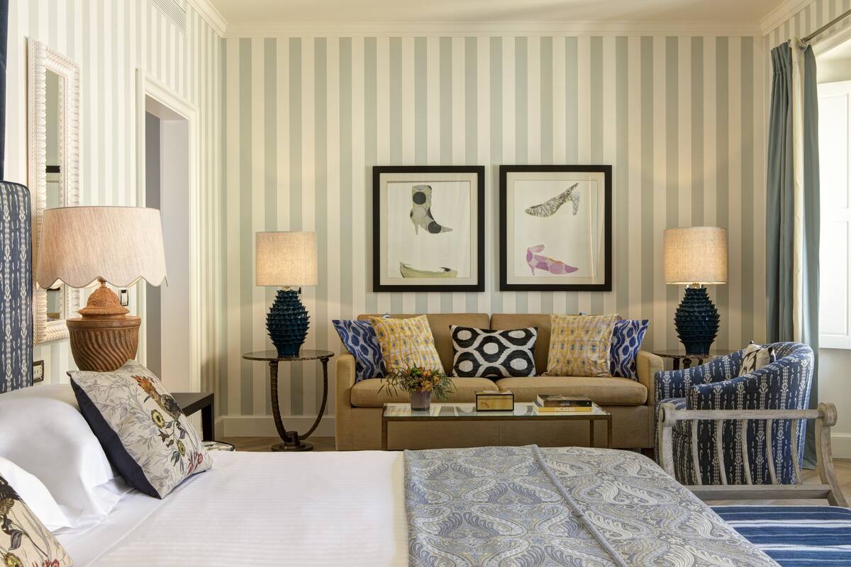 A hotel room with a bed, a sofa, two armchairs, a coffee table, and two lamps. There are striped walls and two framed pictures on the wall.