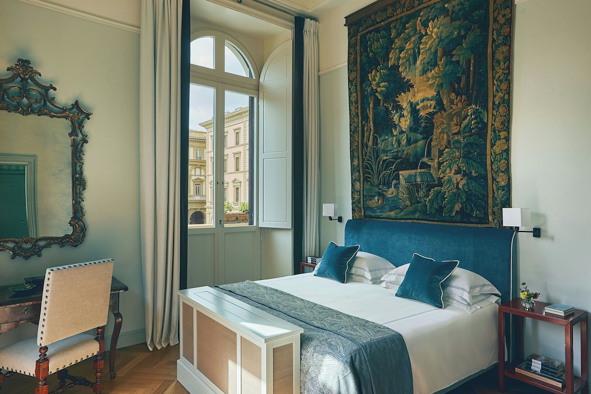 Een hotelkamer met een groot bed, een stoel, een bureau, een raam met gordijnen en een wandtapijt aan de muur.