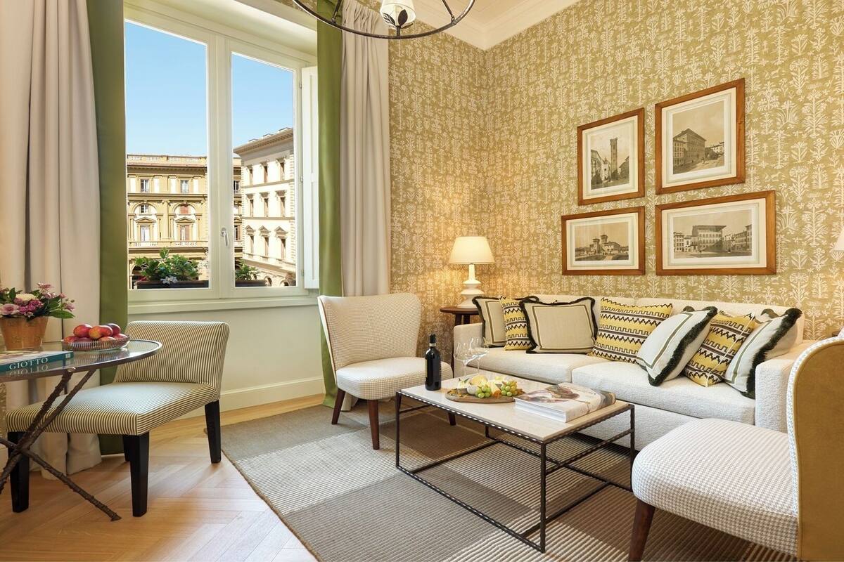 A hotel room with a sofa, armchairs, a coffee table, a side table with a lamp, framed pictures on the wall, and a view of buildings through a window.