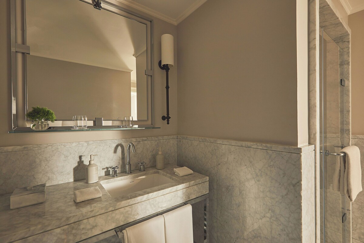 A bathroom with a large mirror, a sink with a marble countertop, a glass shower enclosure, and a towel rack.