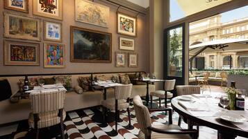A dining area with tables set for two, a variety of framed artworks on the walls, and a view of a building through large windows.