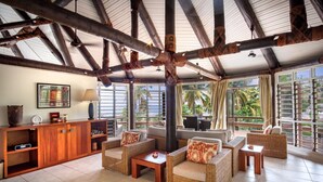 Amplia sala de estar con mobiliario de mimbre, un mueble de madera y grandes ventanas que ofrecen vistas a palmeras.
