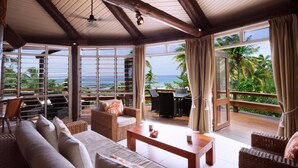 Habitación amplia con un gran ventanal que ofrece vistas al océano y palmeras. Hay un sofá, una mesa y una silla.
