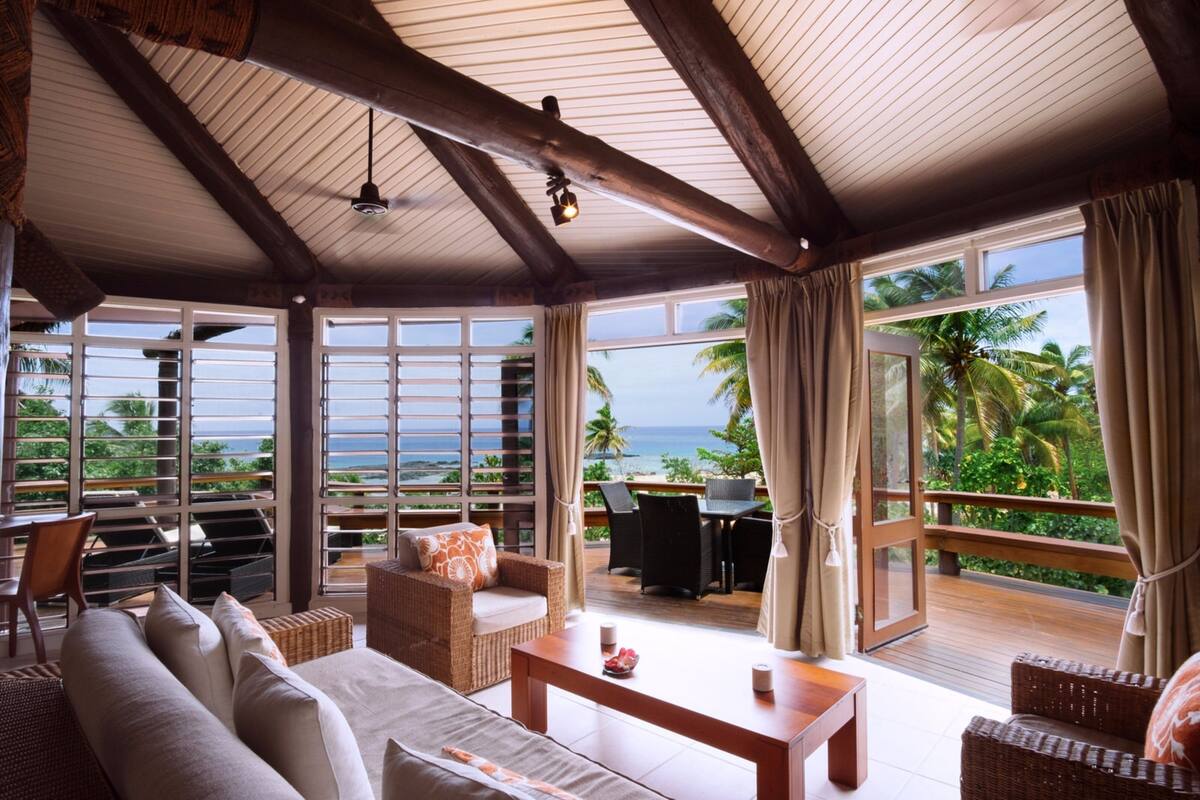 A spacious room with a large window offering a view of the ocean and palm trees. There is a sofa, a table, and a chair.