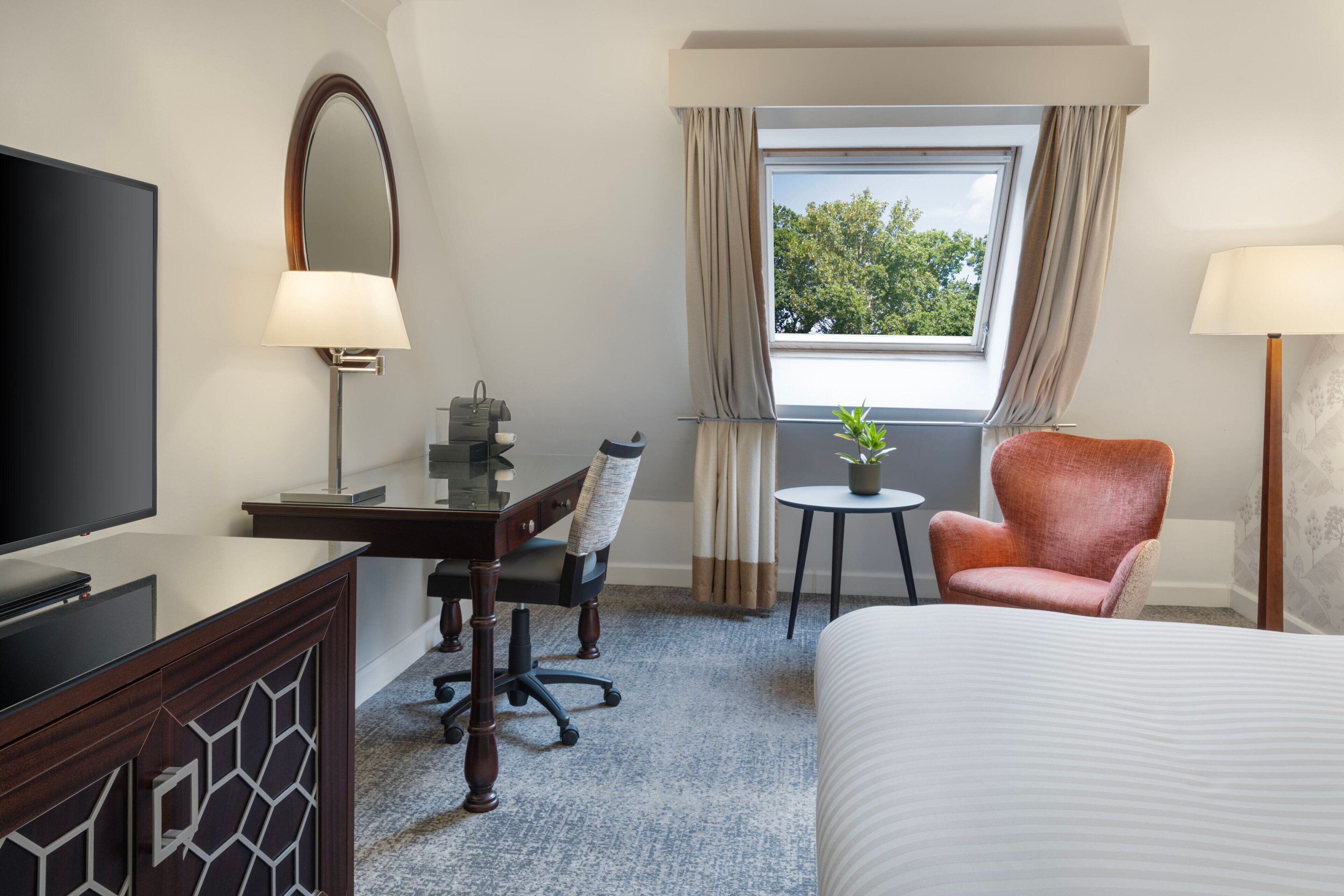 Habitación de hotel con cama, escritorio con silla, televisor, pequeña mesa con planta y ventana con cortinas.