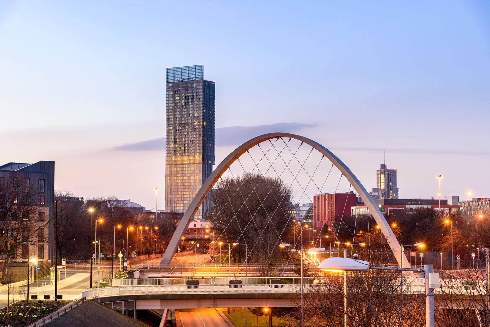 Un puente moderno con una amplia estructura en arco, rodeado de edificios urbanos y farolas.
