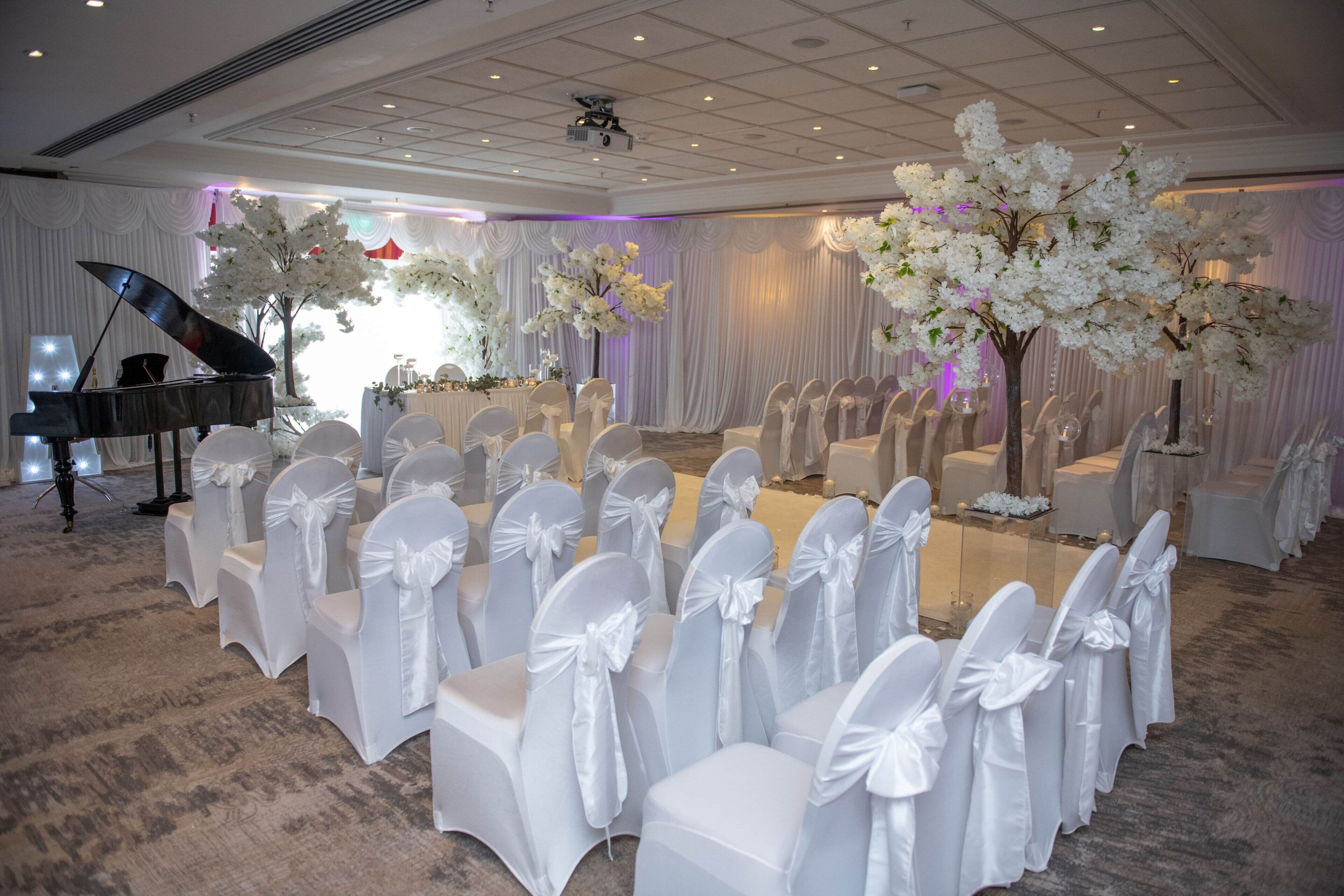 A ballroom with rows of white chairs with bows, a grand piano, and white floral arrangements.