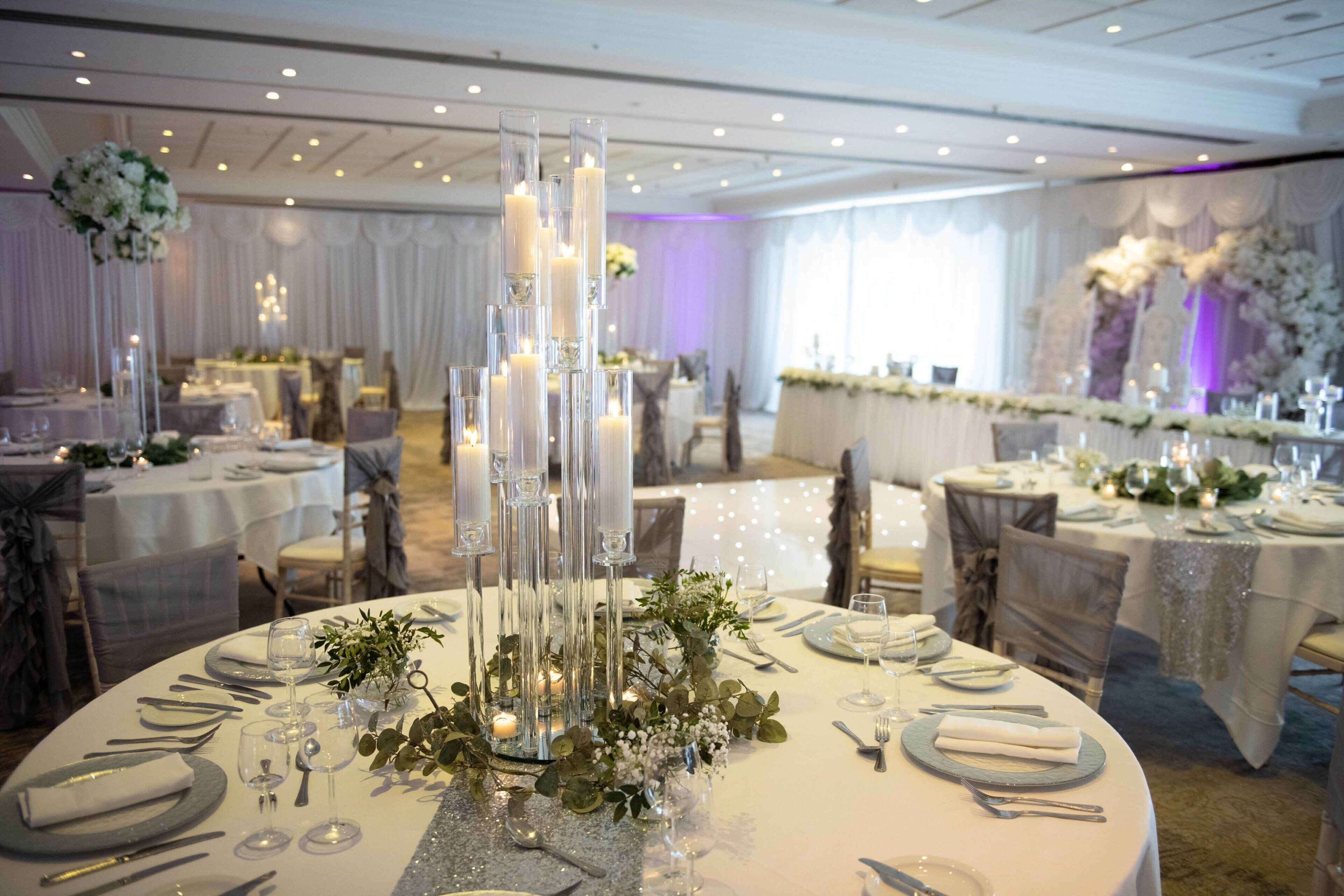 A banquet hall with round tables set for a formal event, featuring tall candle holders, floral centerpieces, and elegant table settings.