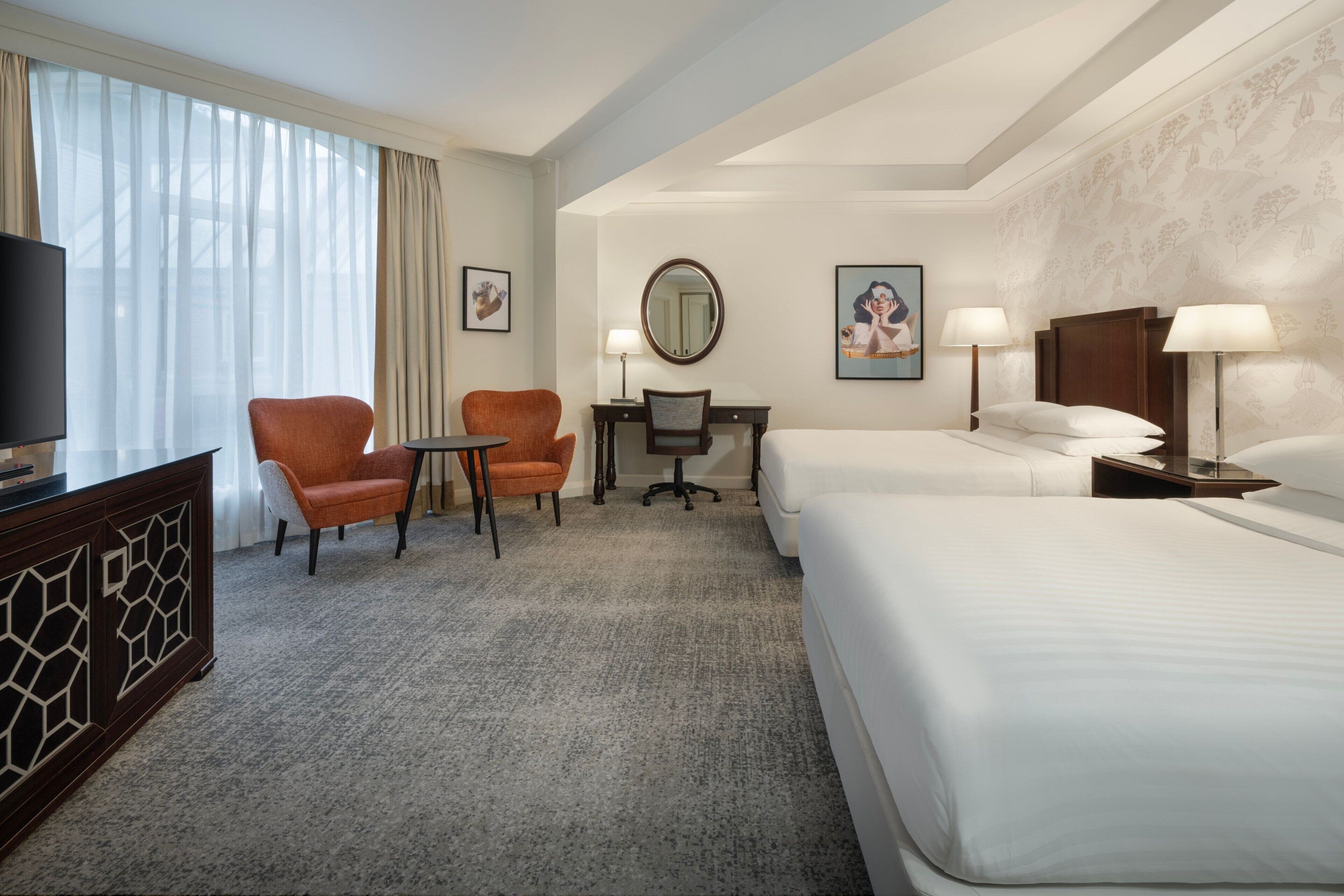 A hotel room with a bed, two chairs, a desk, a TV, and a large window with curtains.