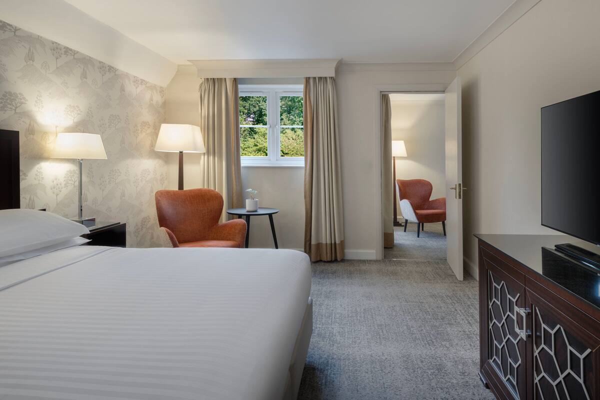 Una habitación de hotel con una cama grande, dos sillas, un televisor y una ventana con cortinas.