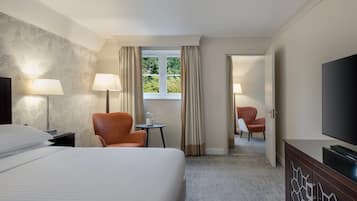 A hotel room with a large bed, two chairs, a TV, and a window with curtains.