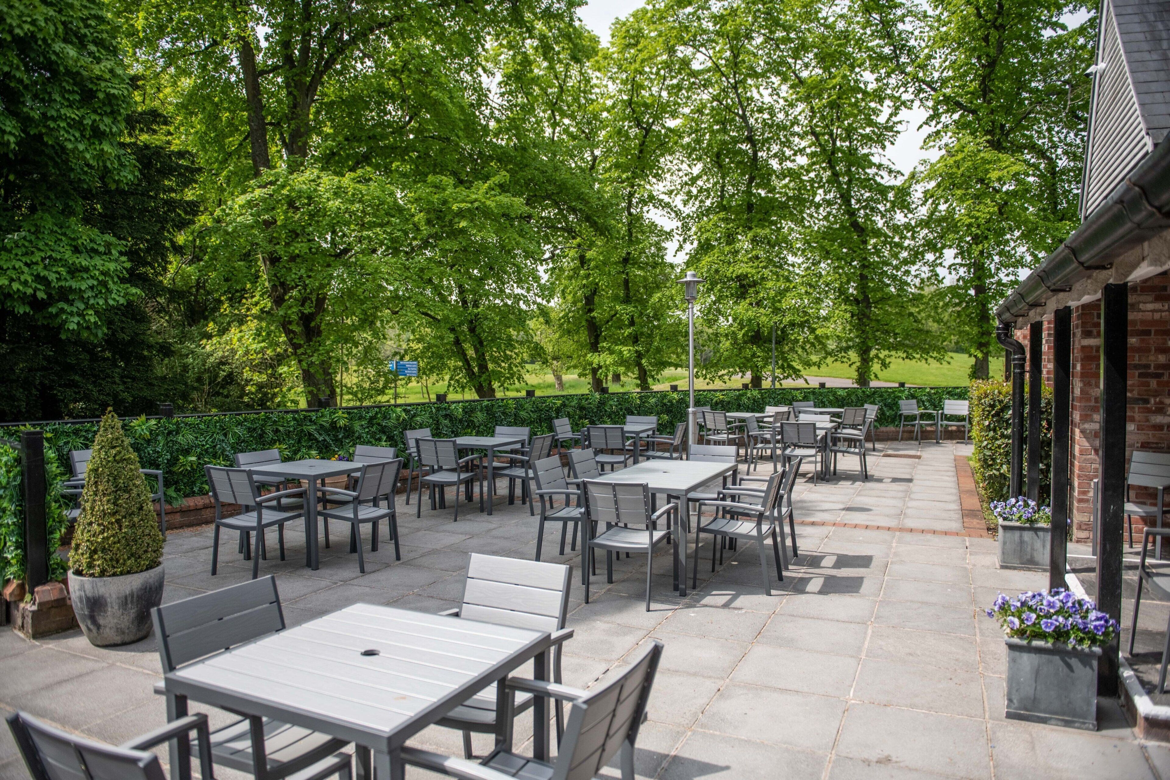 An outdoor patio with tables and chairs, surrounded by greenery and a brick building.