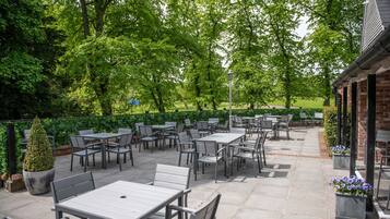An outdoor patio with tables and chairs, surrounded by greenery and a brick building.