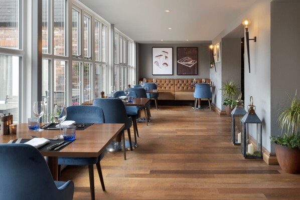 Un comedor moderno con mesas de madera y sillas azules, ventanas amplias y una zona de estar con un banco acolchado.