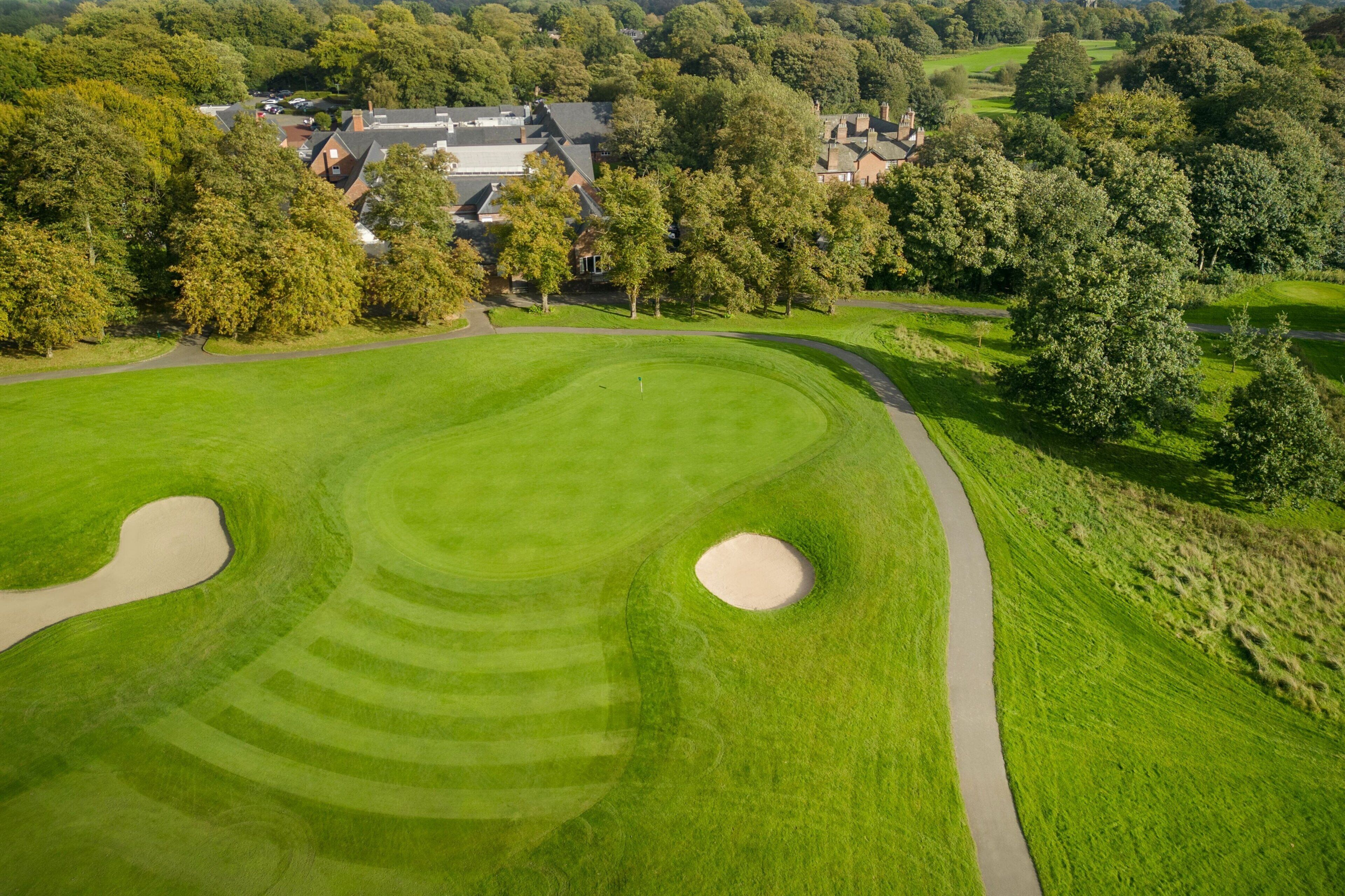 Een golfbaan met zandbunkers, een wandelpad en omliggende bomen.