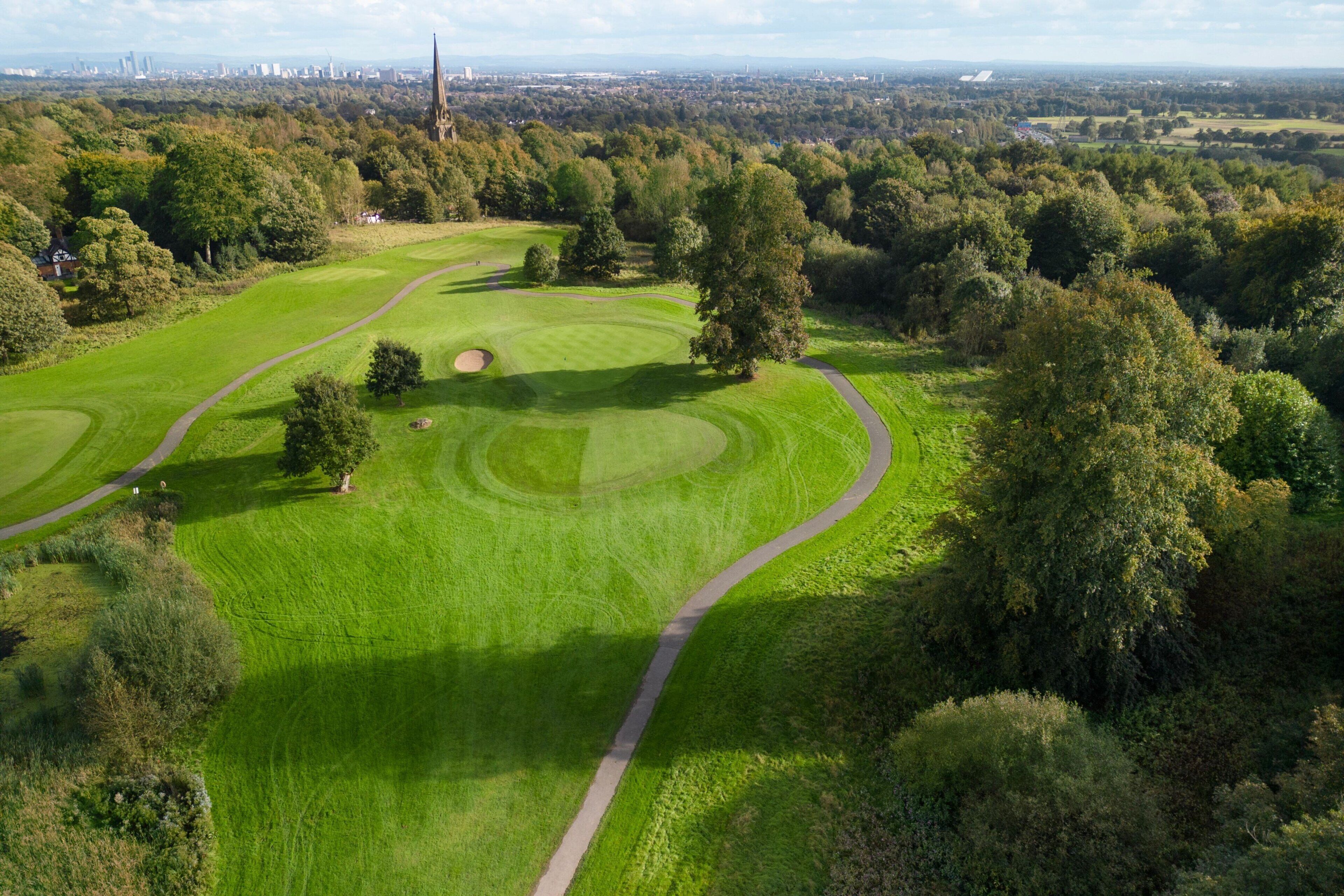 A golf course with well-maintained fairways and greens, surrounded by dense trees and a distant city skyline.