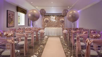 Salón de bodas con un pasillo central, fondo floral y filas de sillas con cintas rosas.