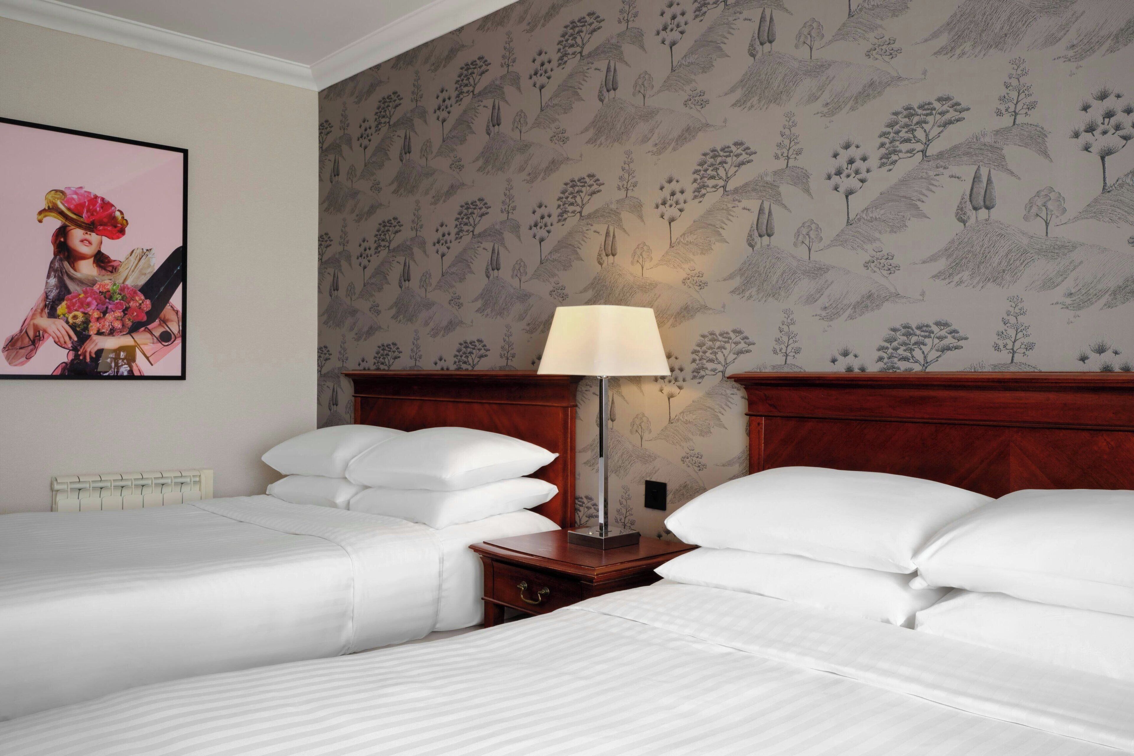 Habitación de hotel con dos camas, una mesita de noche con lámpara y papel tapiz floral.
