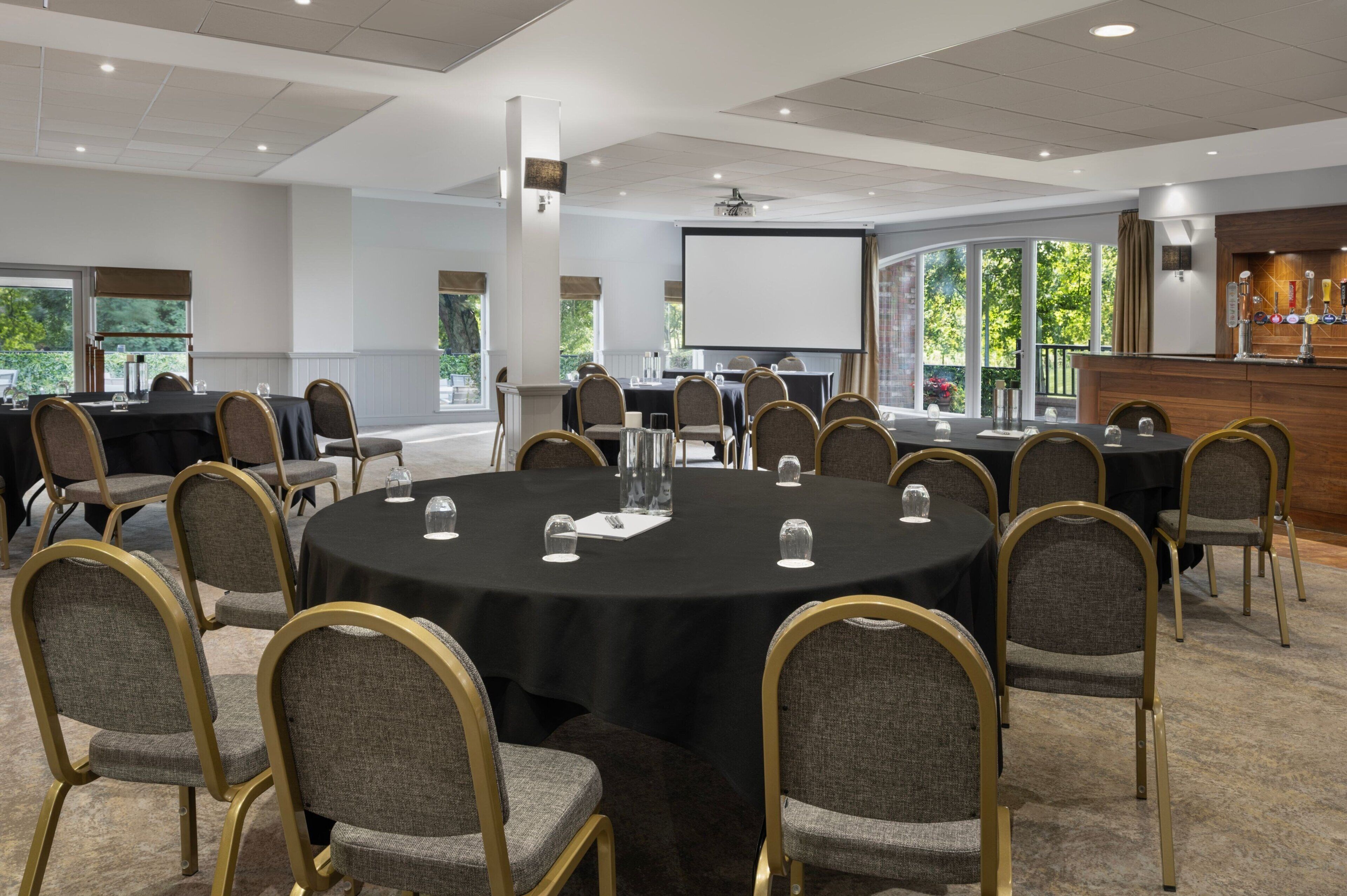 A conference room with round tables, chairs, a projector screen, and a bar area.