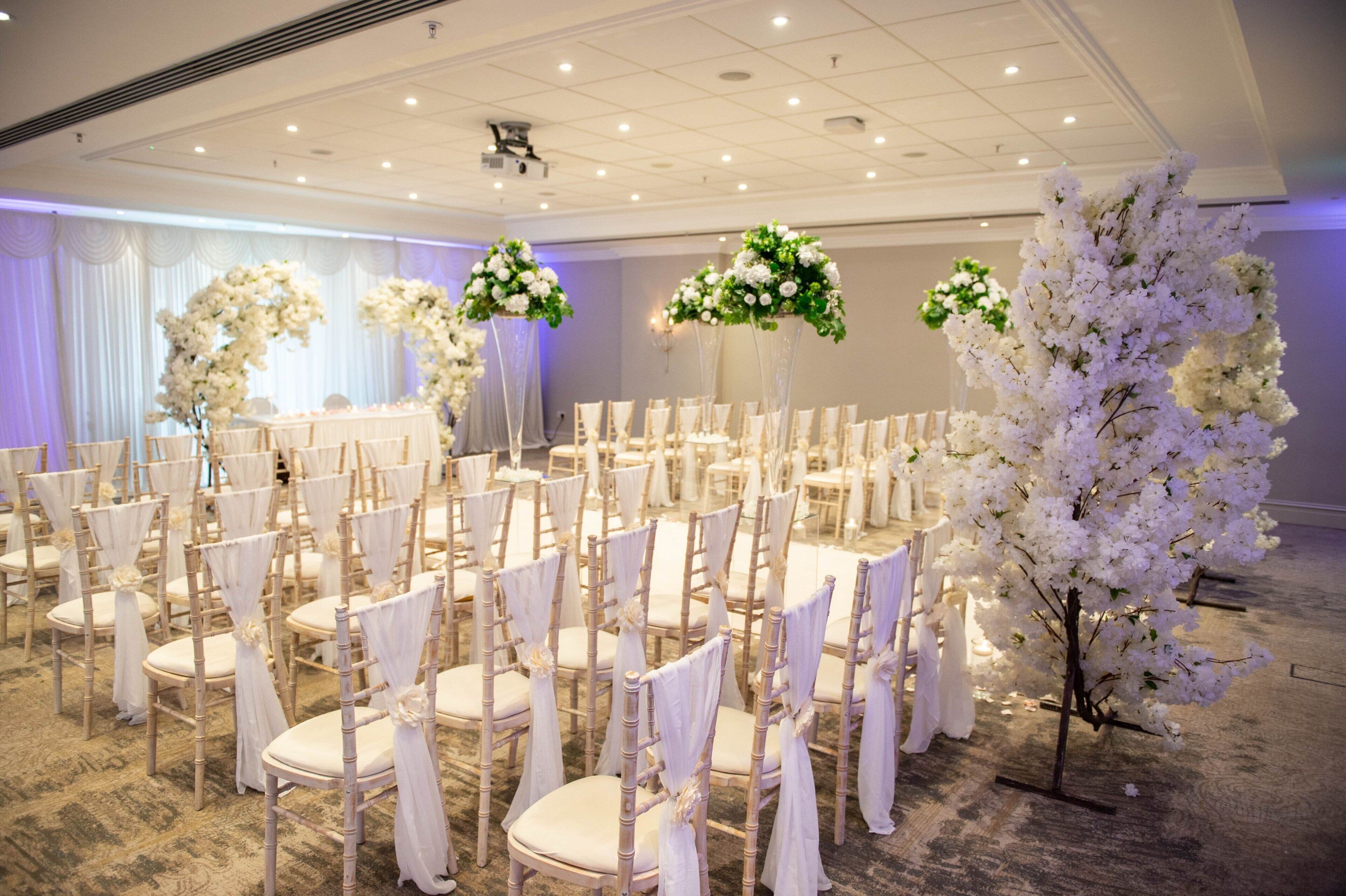 Una location per matrimoni con file di sedie bianche, composizioni floreali al centro dei tavoli e un'ampia decorazione floreale bianca.