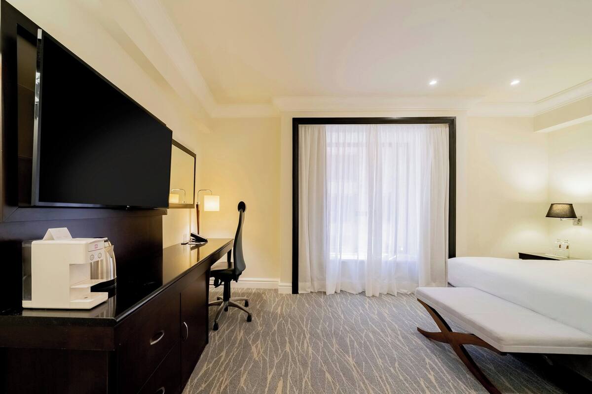 A hotel room with a bed, a desk with a coffee maker, a TV, and a large window with curtains.