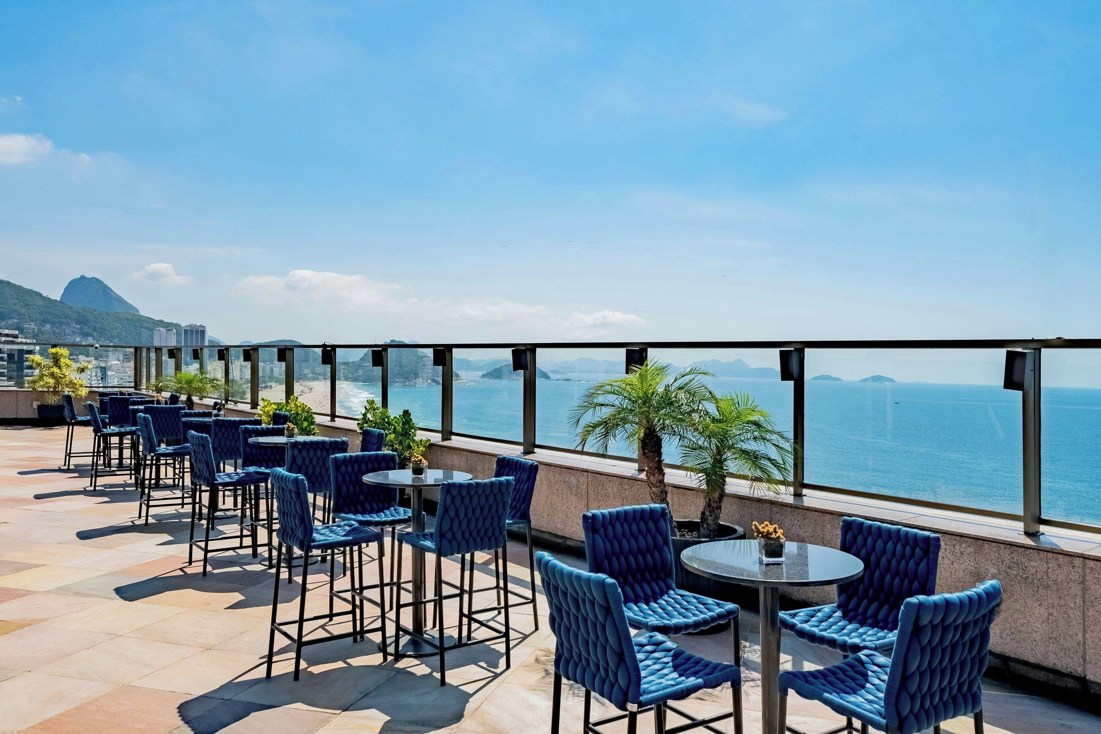 A rooftop terrace with blue upholstered chairs and tables, overlooking a cityscape and the sea.