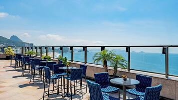 A rooftop terrace with blue upholstered chairs and tables, overlooking a cityscape and the sea.
