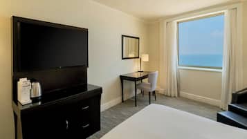 A hotel room with a large bed, a desk with a chair, a flat-screen TV, a coffee maker, and a view of the ocean through a large window.