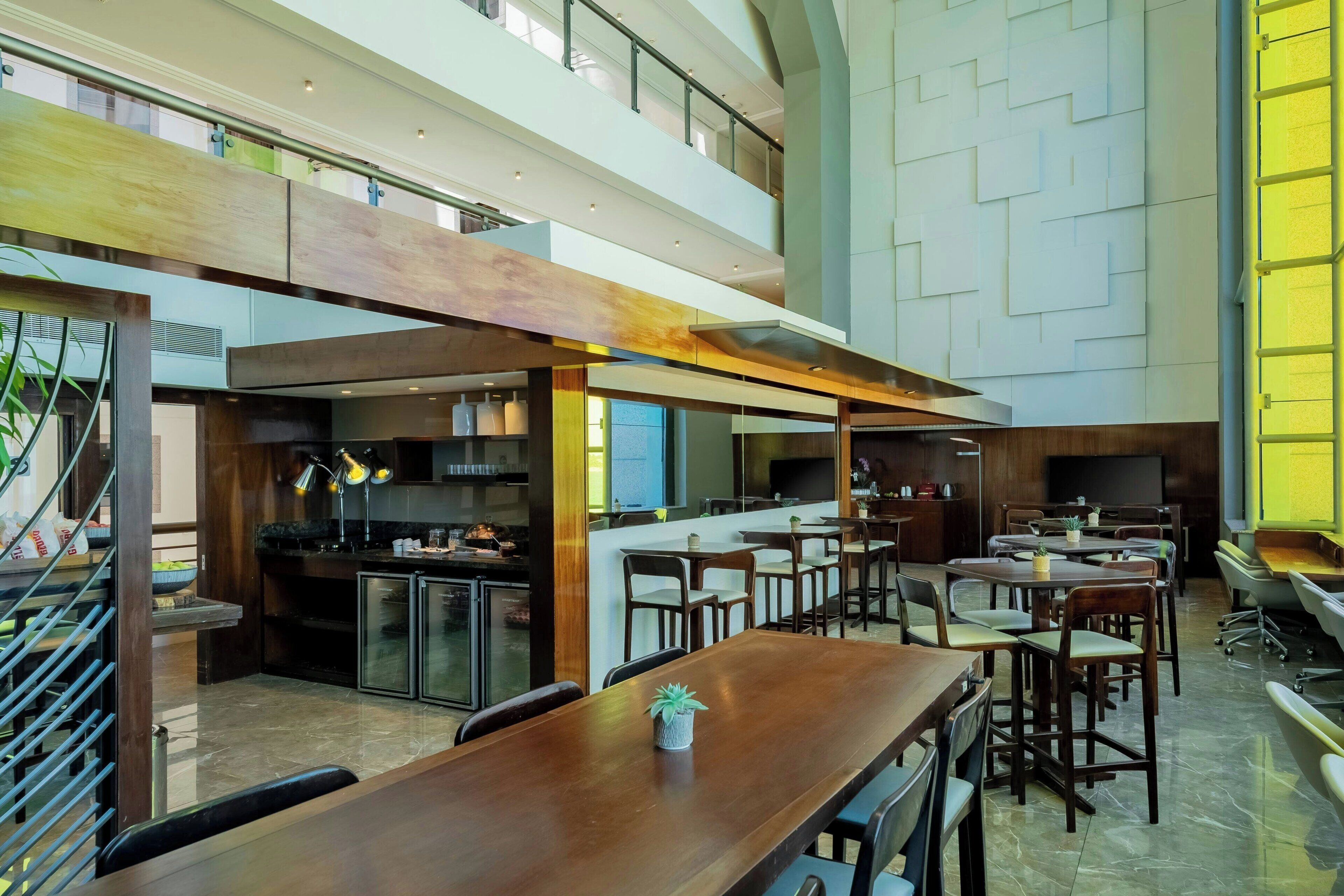A spacious hotel lobby with a high ceiling, multiple dining areas, and a modern kitchen visible in the background.