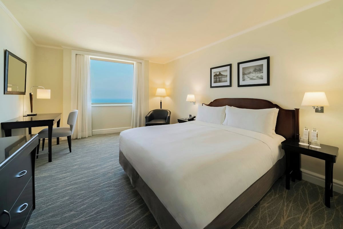 A hotel room with a large bed, a desk with a chair, a small table, two bedside lamps, and two framed pictures on the wall.