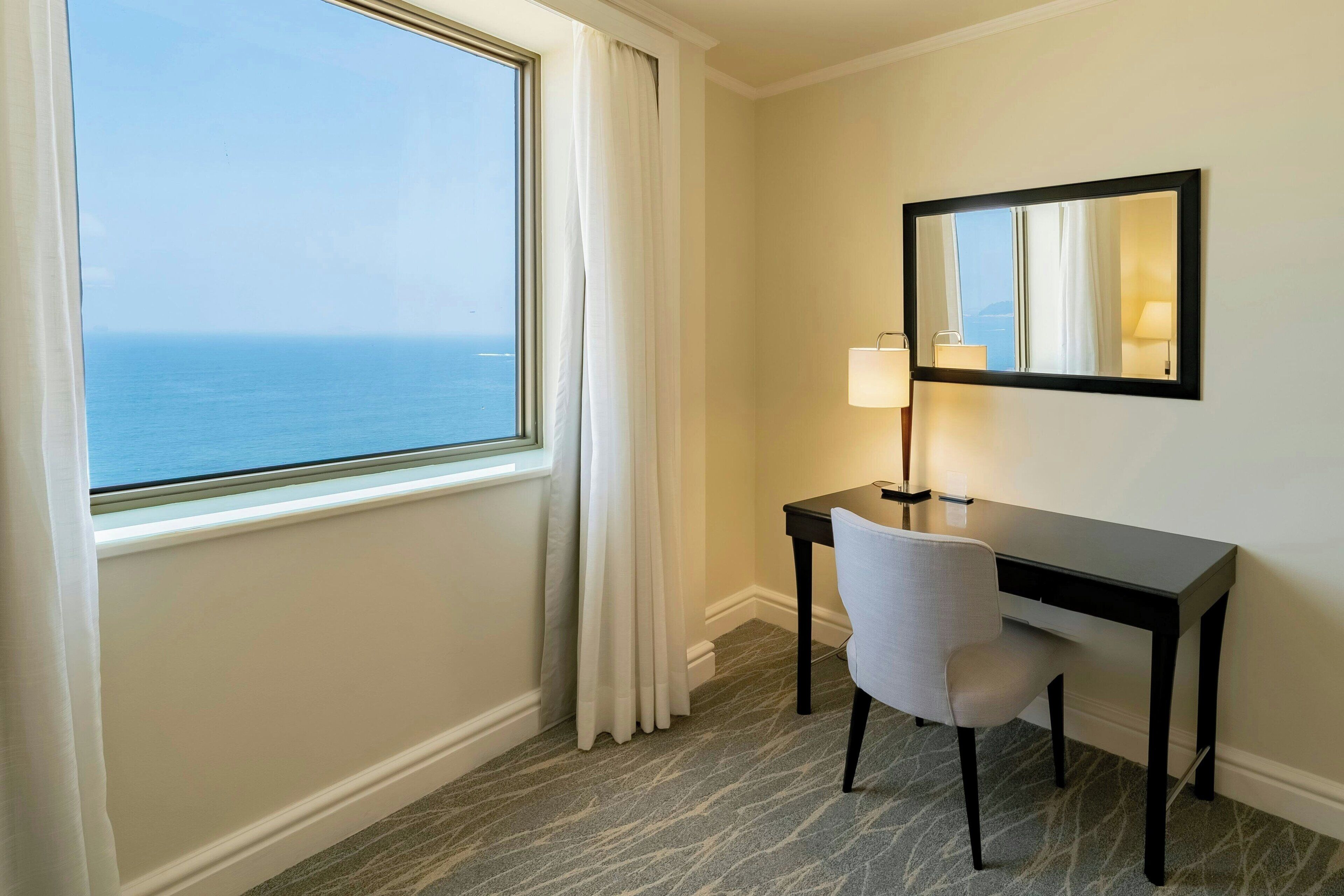 A hotel room with a desk, chair, lamp, and a large window with a view of the ocean.
