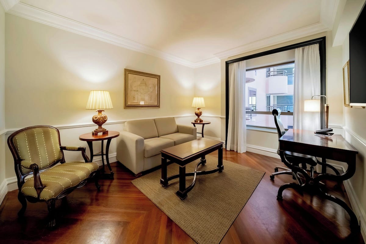A living room with a sofa, armchair, coffee table, and a dining table with chairs. There are two lamps on side tables, a painting on the wall, and a view through a window.