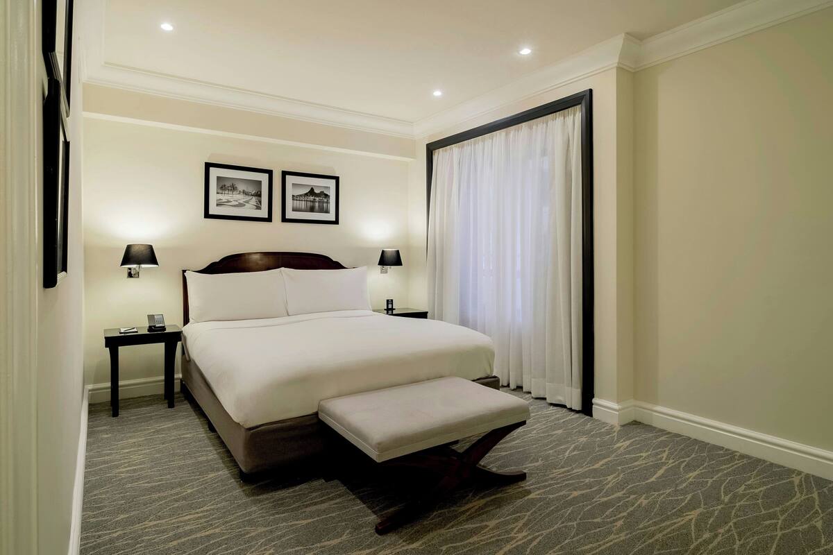 A hotel room with a large bed, two bedside tables with lamps, a bench at the foot of the bed, and two framed pictures on the wall.