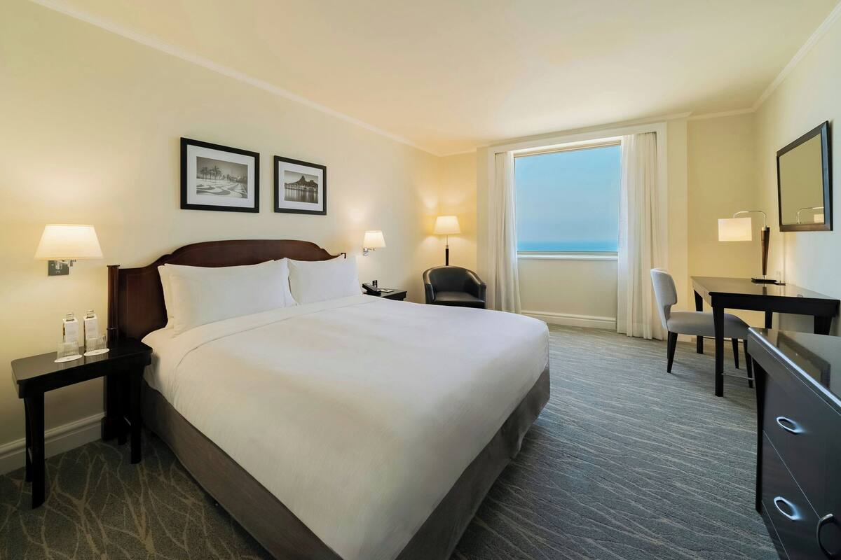 A hotel room with a large bed, two bedside tables with lamps, a desk with a chair, a large window with curtains, and two framed pictures on the wall.