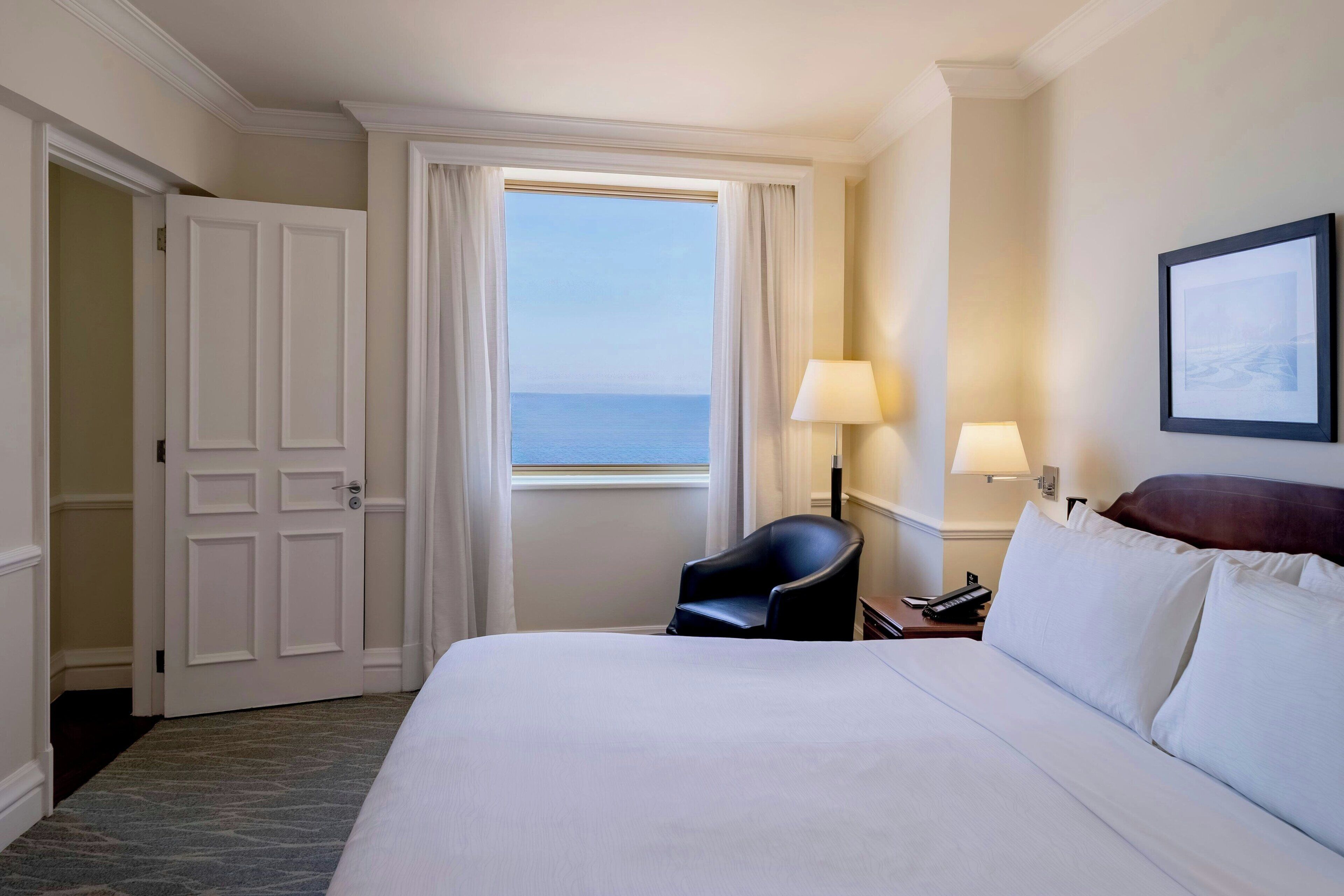 A hotel room with a large bed, a chair, a desk with a lamp, a framed picture on the wall, and a window with curtains.