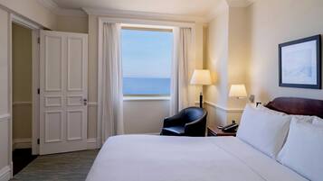 A hotel room with a large bed, a chair, a desk with a lamp, a framed picture on the wall, and a window with curtains.