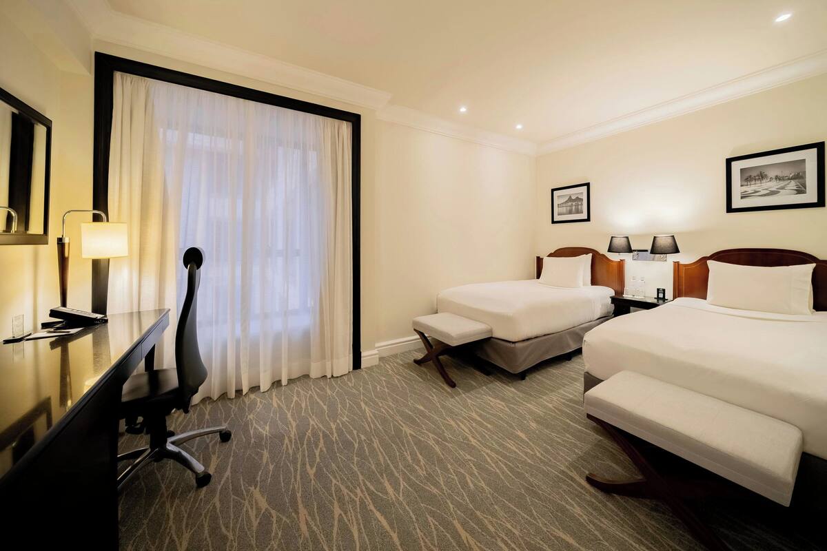 A hotel room with two beds, a desk, a chair, and a large window with curtains.