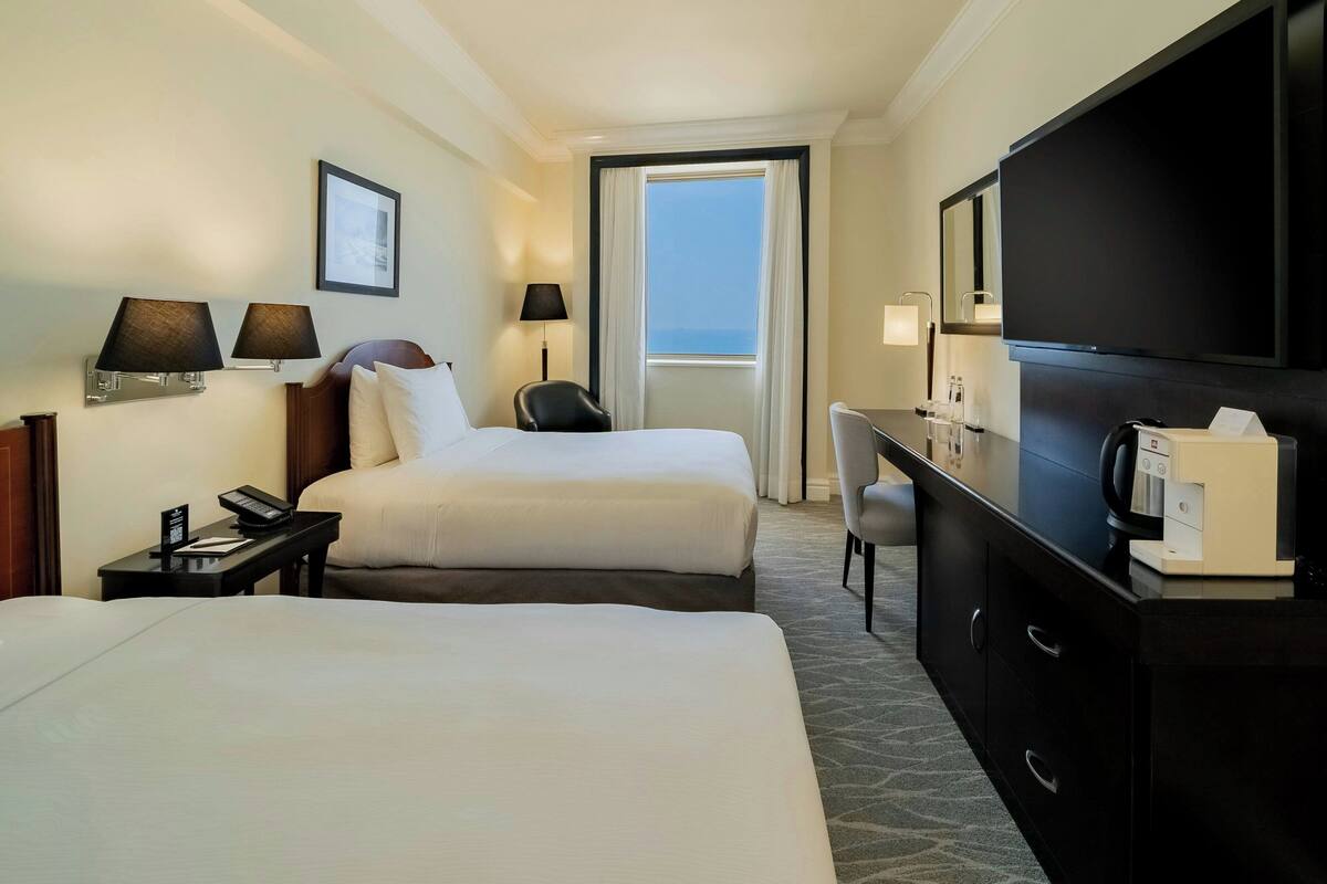 A hotel room with two beds, a desk, a chair, a TV, and a window with a view.