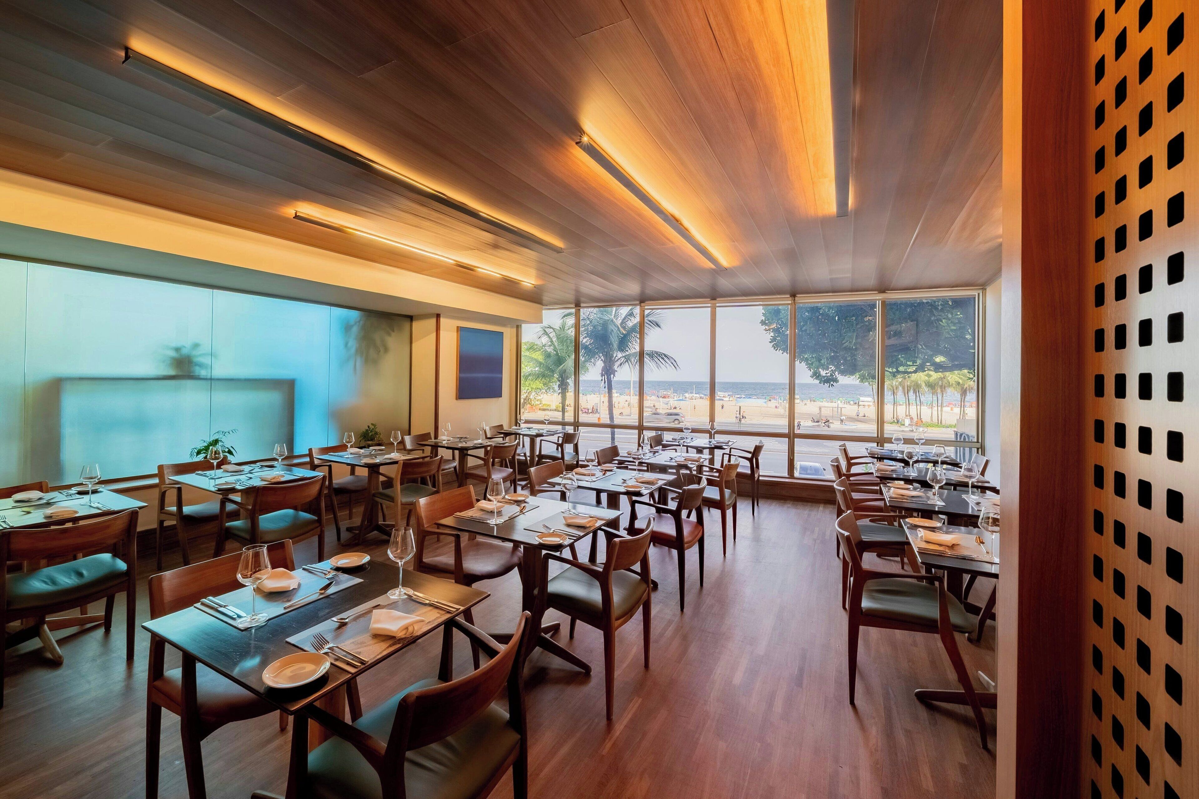 A restaurant with wooden floors, tables set for dining, and large windows offering a view of palm trees and a beach.