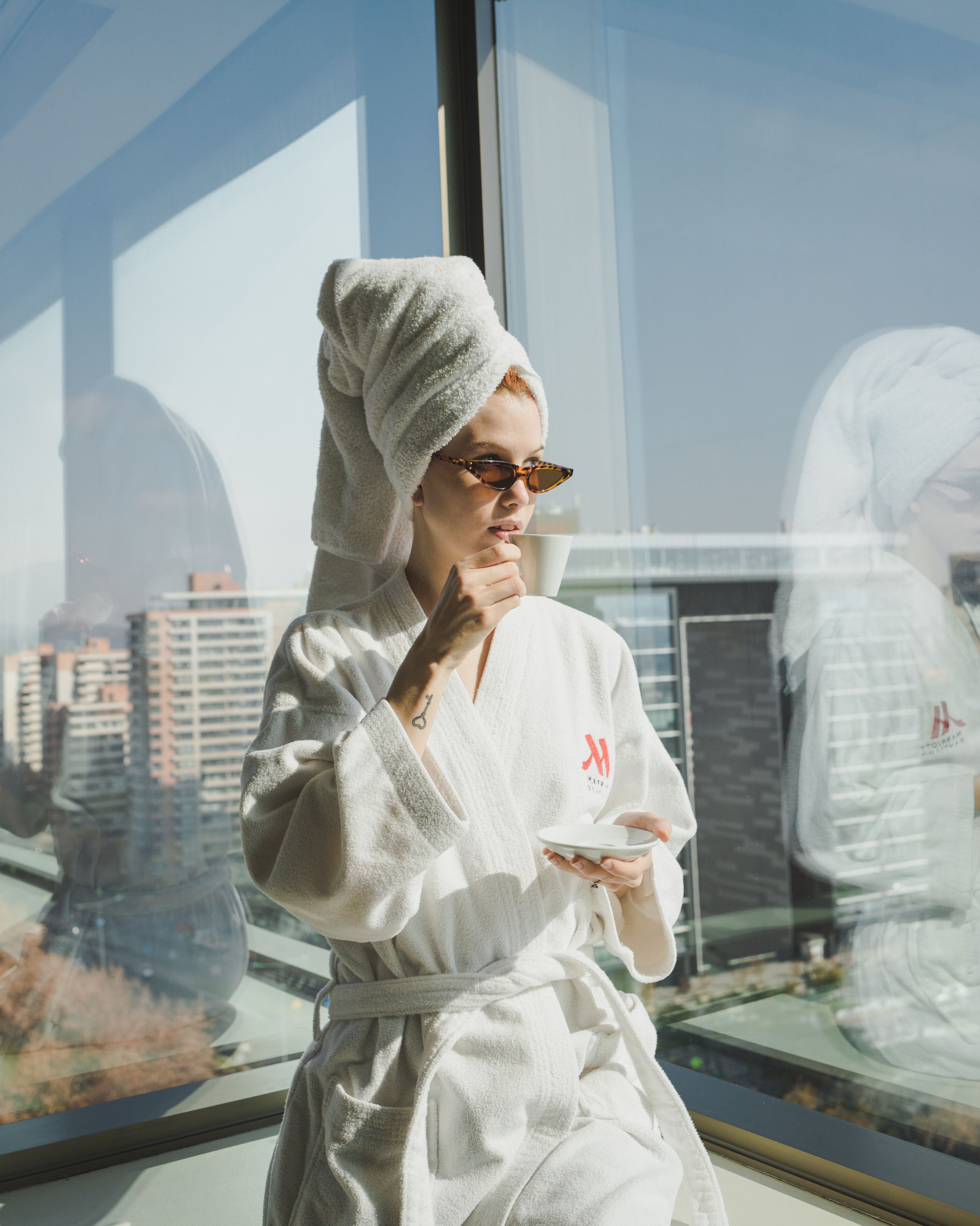 A person in a white robe with a towel on their head, holding a cup, standing by a window with a cityscape view.