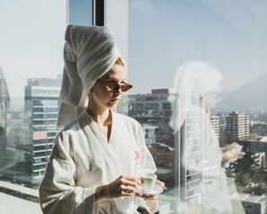 A person in a white robe with a towel on their head, holding a cup, standing by a window with a cityscape view. - Santiago Marriott Hotel (Santiago)