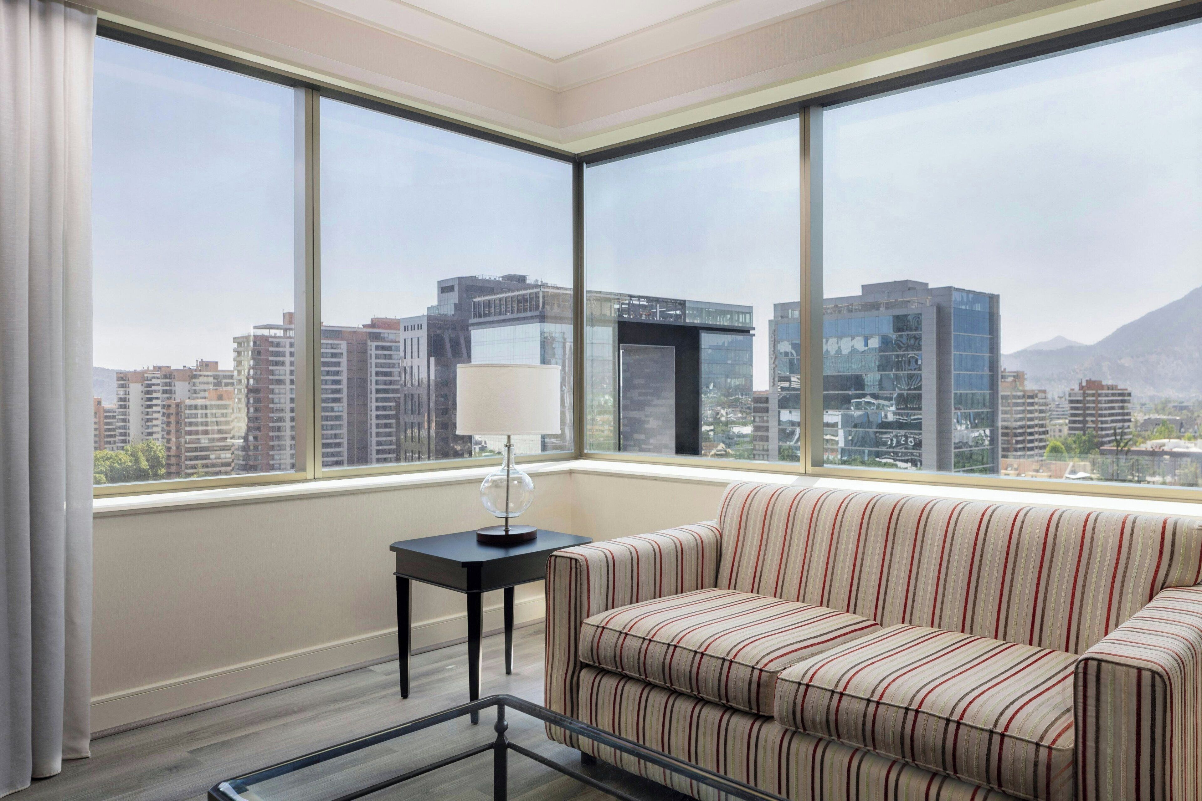 Ein modernes Hotelzimmer mit großem Fenster und Blick auf die Stadt, einer gestreiften Couch, einem kleinen Tisch mit Lampe sowie einem Glastisch.