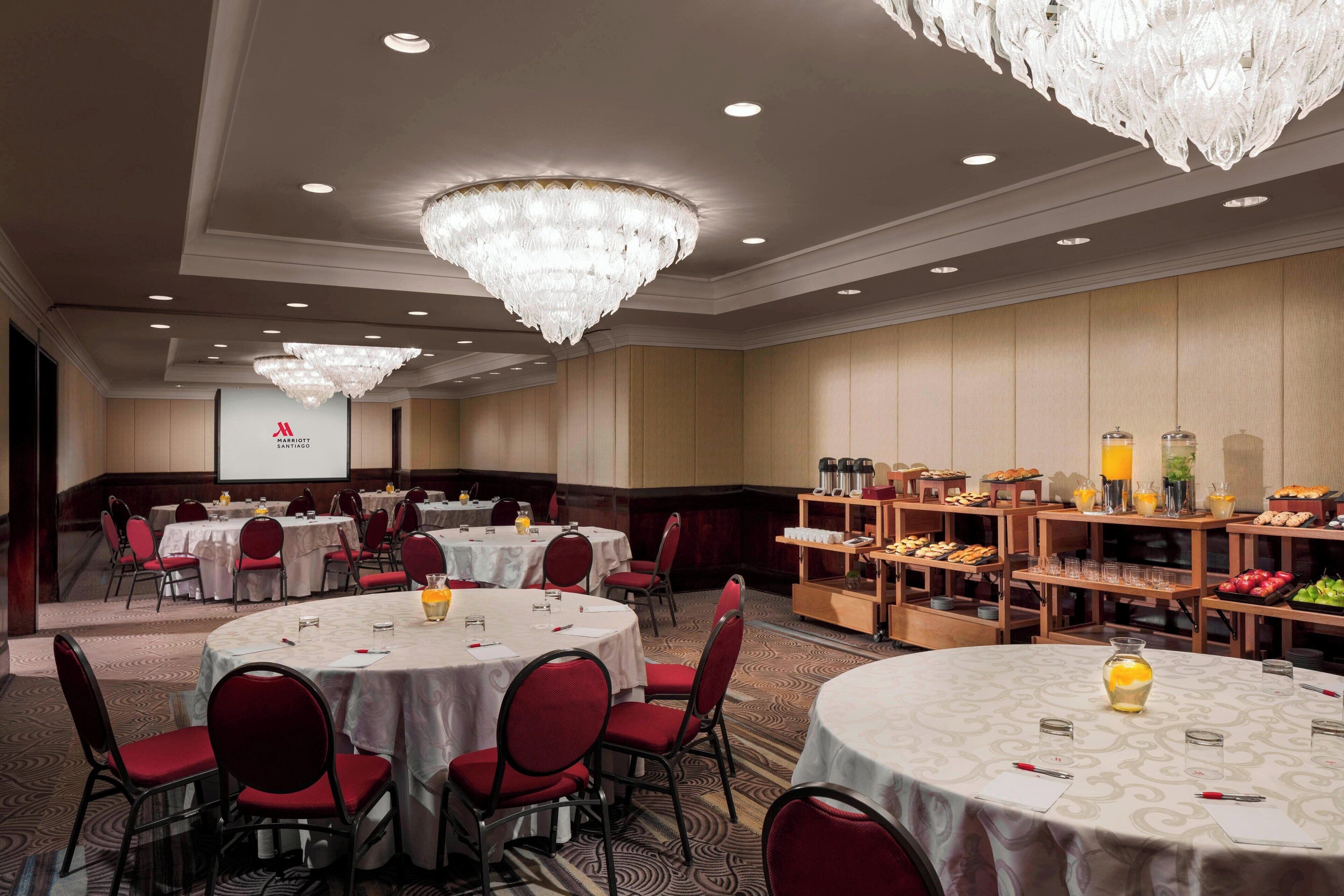 Une salle de banquet avec des tables rondes, des chaises rouges, des lustres et un espace buffet proposant des plats et des boissons.