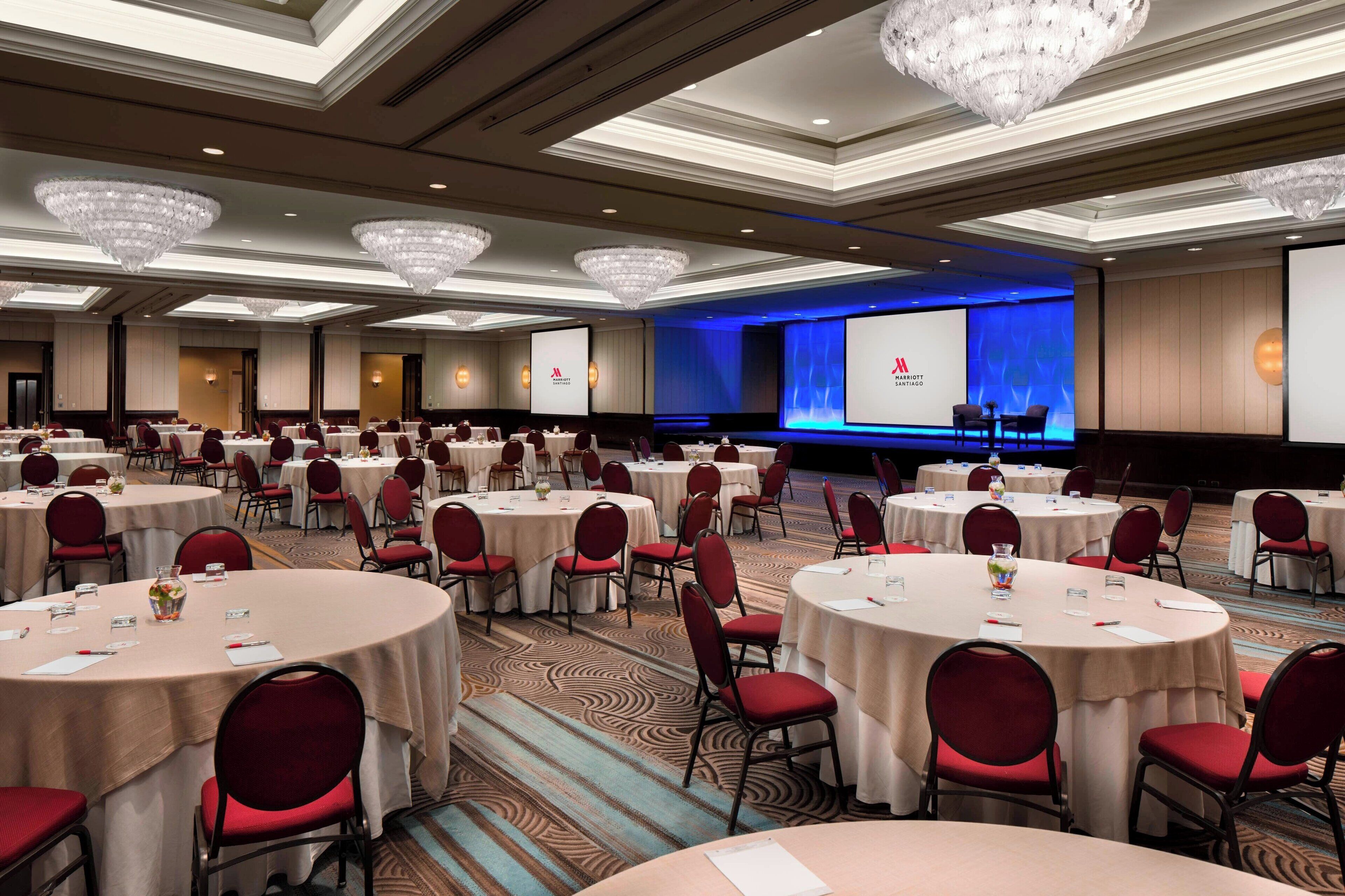 Un salón de conferencias con varias mesas redondas, sillas rojas y un escenario con una pantalla de presentación.