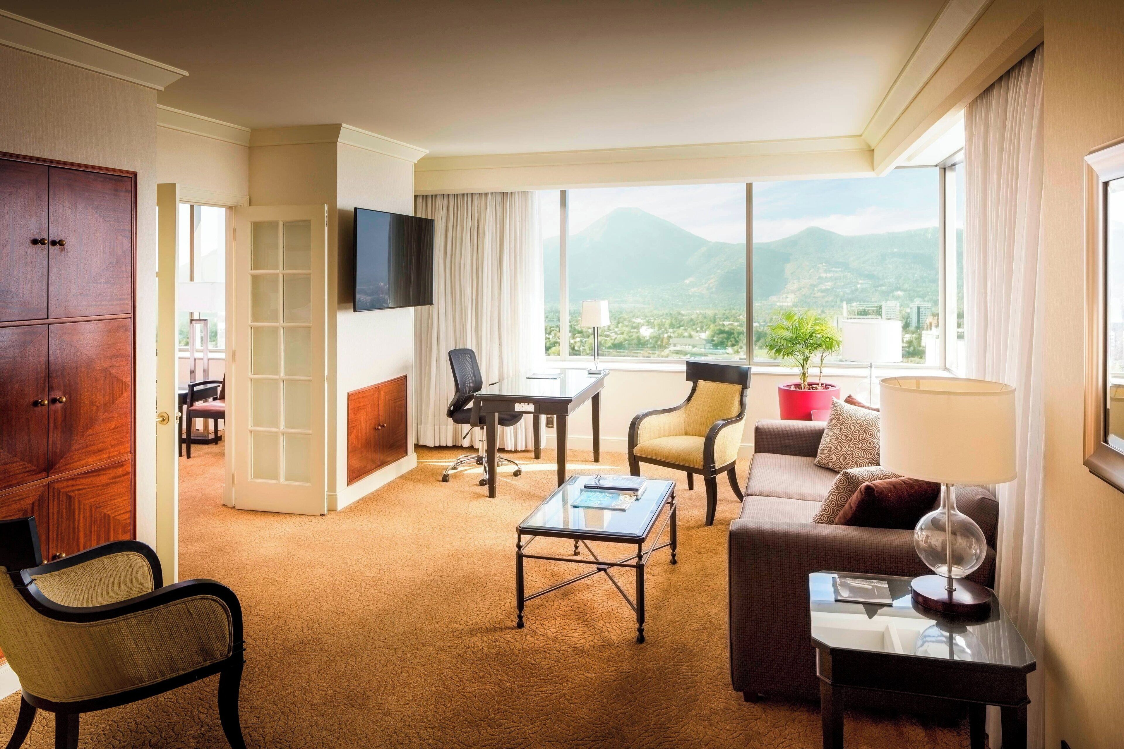 Camera d'albergo con divano, poltrona, tavolino, televisore e vista sulle montagne.