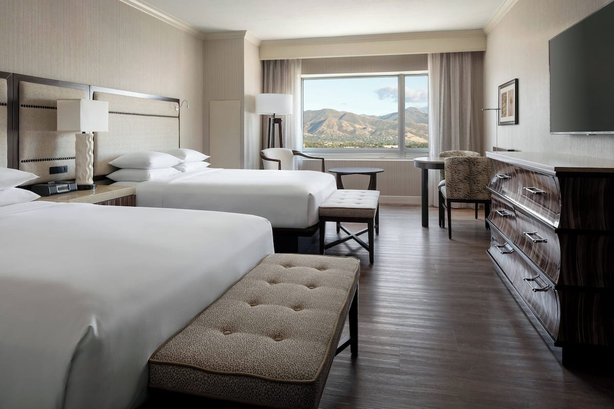 A hotel room with two beds, a desk, a chair, a TV, and a view of mountains.