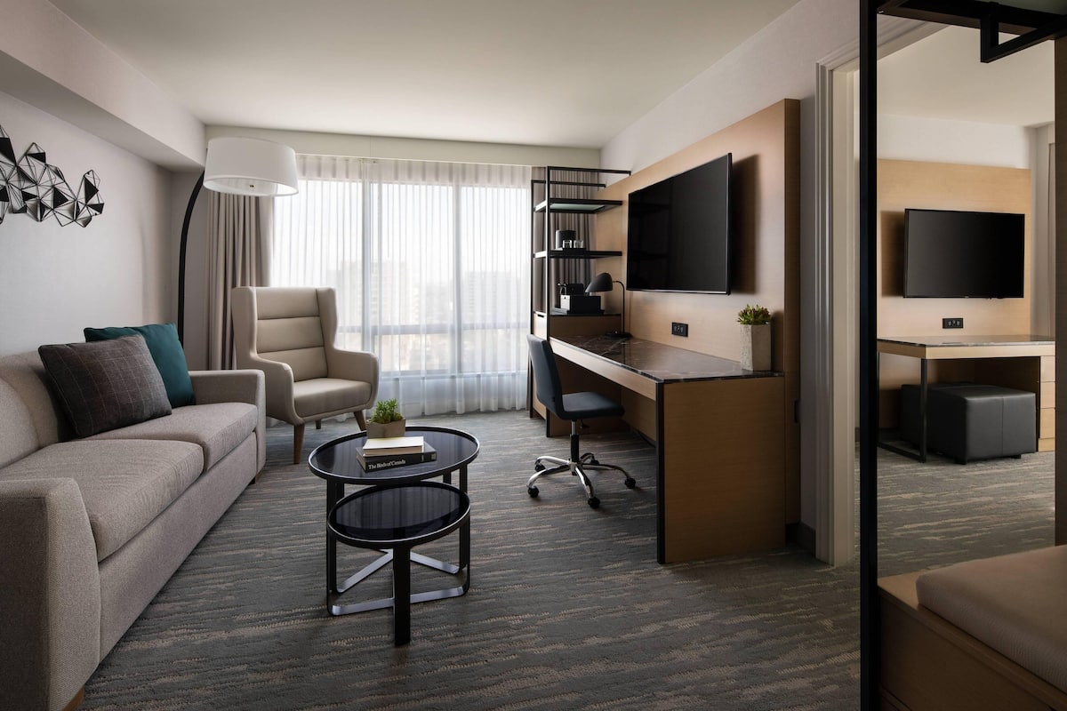Una habitación de hotel moderna con sofá, escritorio, silla, televisor y una ventana grande con cortinas traslúcidas.
