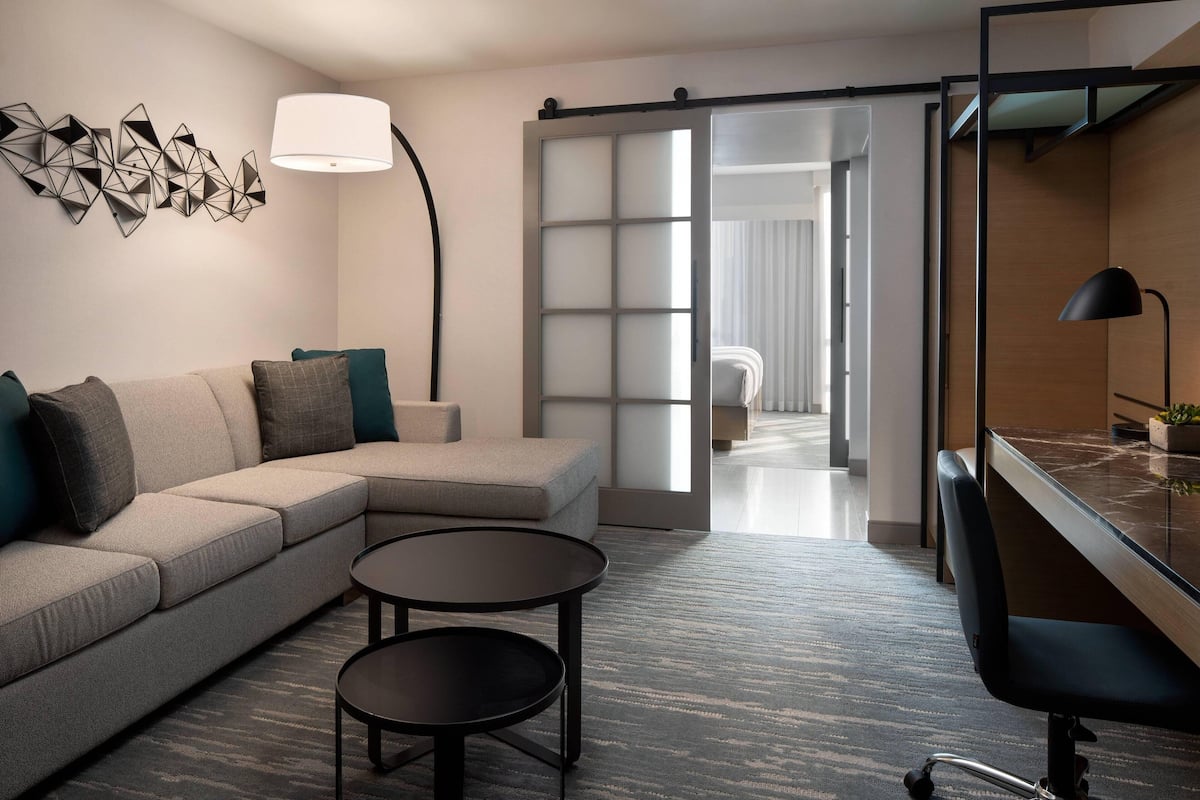 Una habitación de hotel moderna con un sofá, una mesita redonda pequeña, un escritorio con una silla, una lámpara y una obra de arte geométrica en la pared.